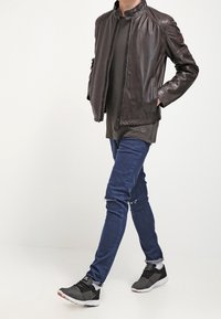 Brun läderjacka med hög krage, dragkedjestängning och sidofickor. Bärs över en mörk långärmad skjorta, tillsammans med blå jeans och svarta sneakers.