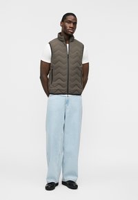 Moški stoji z rokami za hrbtom, oblečen v rjavo prešito telovnik čez belo majico, svetlo modre široke kavbojke in črne čevlje na belem ozadju.