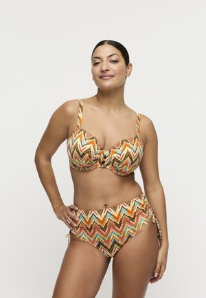 Vrouw poseert zelfverzekerd in een kleurrijke bikini met een zigzagpatroon, hoge taille en verstelbare schouderbandjes, tegen een effen achtergrond.