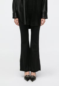 Camicia lunga in pelle nera su pantaloni neri aderenti con spacchi laterali, abbinata a décolleté nere con dettagli dorati. Texture liscia.