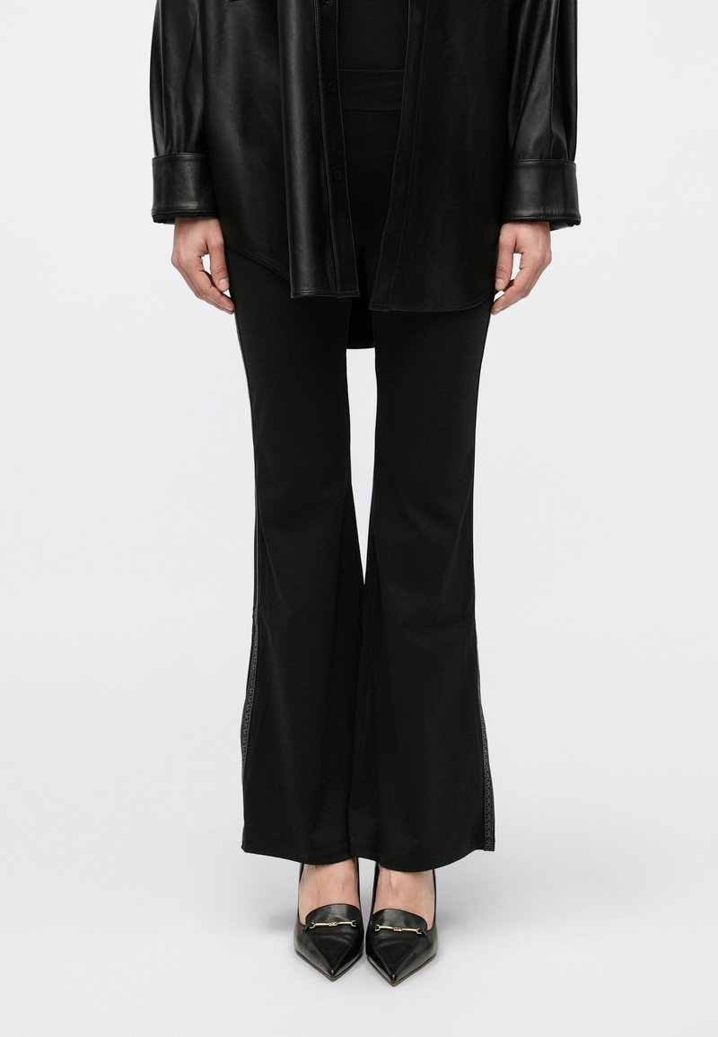 Camicia lunga in pelle nera su pantaloni neri aderenti con spacchi laterali, abbinata a décolleté nere con dettagli dorati. Texture liscia.