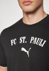 Černé bavlněné tričko s bílým nápisem "FC ST. PAULI" a skákajícím logem pumy. Klasický kulatý výstřih a krátké rukávy.