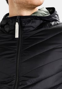 Svart vadderad jacka med ett chevron-quiltat mönster, figursydd huva och dragkedja framtill. Inuti finns en kontrasterande grön foder.