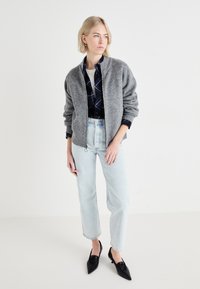Veste bomber grise en tricot avec texture côtelée, chemise à carreaux noire en dessous, jeans bleu clair et ballerines noires à bout pointu.