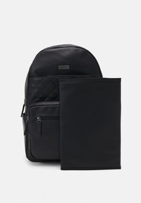 Zwarte rugzak van glad materiaal met een gewatteerd patroon bovenop, voorzien van een voorvak met rits en een afneembare zwarte hoes.