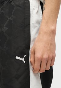 Κοντινό πλάνο ενός χεριού που ακουμπά δίπλα σε αθλητικό σορτς Puma σε ασπρόμαυρο, με διακριτικό μοτίβο και λευκό λογότυπο Puma.