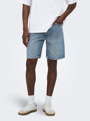 Personne portant un short en jean bleu clair, des chaussettes blanches, des baskets blanches et un T-shirt blanc avec une main dans la poche.