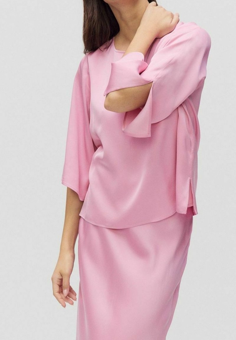 Femme en chemisier en satin rose clair avec des découpes aux coudes et une jupe assortie, posant avec une main sur le cou et l'autre détendue le long du corps.