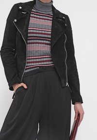 Svart mocka bikerjacka med silverdragkedjor, bärs över en randig polotröja i röd och grå. Löst svarta byxor och håller en handväska.