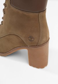 Bottine en daim marron avec un design incurvé, dotée d'un talon empilé, d'accents en cuir texturé, et d'un logo estampé sur le talon.
