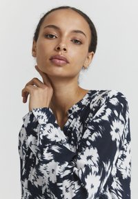 Blouse motif marine et blanche avec des manches longues, col en V et tissu texturé. Présente un design abstrait subtil et des détails de boutons au niveau du col.