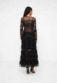 Black lace dress with long sleeves, sheer layers, and ruffled hem. Features a keyhole back and ankle-length design. Worn with black heels.