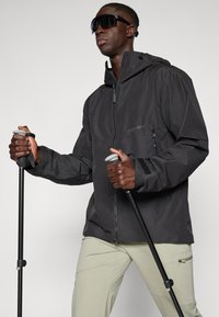 Veste noire imperméable avec poches zippées et capuchon ajustable, accompagnée de pantalons de couleur claire. L'utilisateur tient des bâtons de trekking.