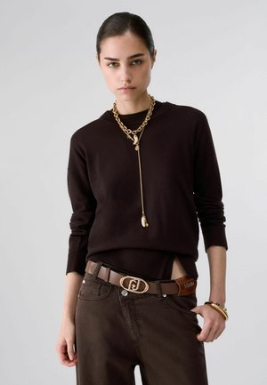 Giovane donna con un maglione marrone scuro, collana a catena dorata, cintura abbinata e braccialetti su uno sfondo grigio uniforme.