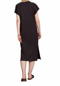Femme portant une robe midi noire à manches courtes avec une fente sur le côté droit à l'arrière, des sandales noires et des bracelets dorés au poignet gauche.