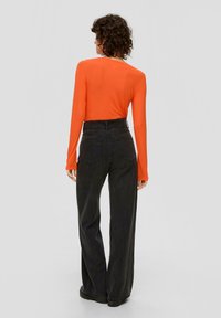 Langärmliges orangenes Oberteil kombiniert mit hochgeschnittenen schwarzen Jeans. Das Oberteil hat einen runden Halsausschnitt; die Jeans haben einen geraden Beinverlauf und Gesäßtaschen.