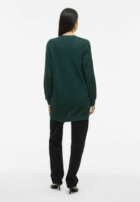 Dolge temno zelene pletene pulover z teksturiranim vzorcem in rebrastimi manšetami. Ujeman s črnimi hlačami in črnimi čevlji na peto.