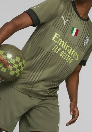 Maglia da soccer verde oliva con accenti neri, caratterizzata da strisce verticali, logo "Emirates" e bandiera italiana. Il giocatore tiene in mano un pallone da calcio a motivi.