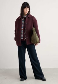 Bordeaux, oversize wełniany płaszcz, sweter w wzory, szerokie ciemnoniebieskie dżinsy oraz zielona torebka ze zamszu o gładkiej fakturze. Czarne buty.