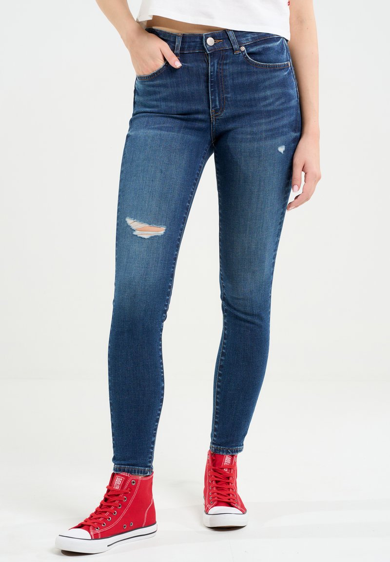 Augstās jostas vietas zili džinsi ar šauru piegriezumu, ar plēstu dizainu uz kreisā augšstilba. Saskaņoti ar spilgti sarkaniem augstiem čības.