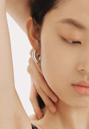 Pendientes curvados de plata exhibidos en la oreja de un modelo, que muestran un acabado suave y brillante, así como un diseño moderno y minimalista, sobre un fondo neutro.