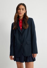 Granatowa marynarka dwuczęściowa z klapami, z dwoma guzikami i dwiema przednimi kieszeniami. Połączona z czerwoną koszulą z kołnierzykiem oraz plisowaną granatową spódnicą.