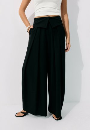 Femme portant un pantalon noir taille haute à jambes larges avec un détail de ceinture pliée, un crop top blanc sans manches, des tongs noires et des bracelets au poignet gauche.