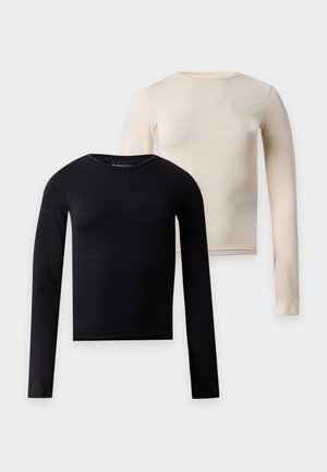 Femme aux cheveux bruns raides portant un haut crème à manches longues, un pantalon large en velours noir et des baskets basses noires, debout les mains dans les poches.