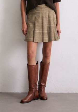 Jupe à carreaux beige avec des plis, associée à des bottes en cuir marron hautes avec des zips et des sangles. Texture lisse et design ajusté.