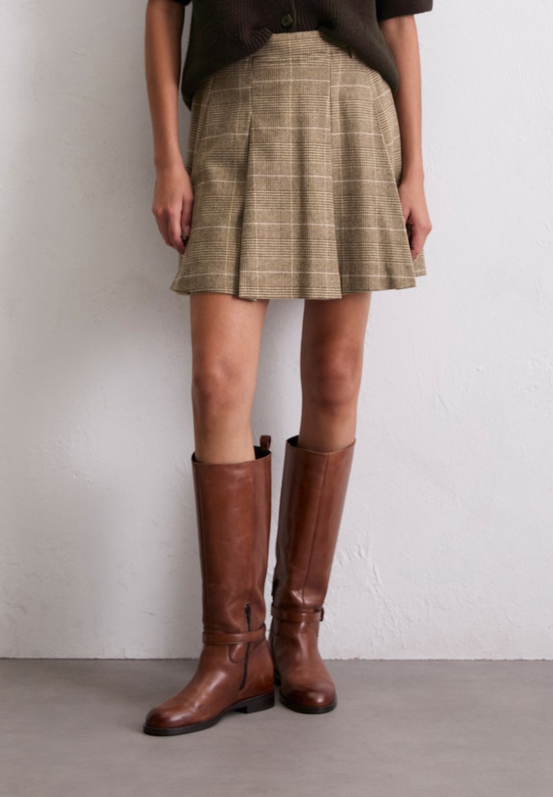 Jupe à carreaux beige avec des plis, associée à des bottes en cuir marron hautes avec des zips et des sangles. Texture lisse et design ajusté.
