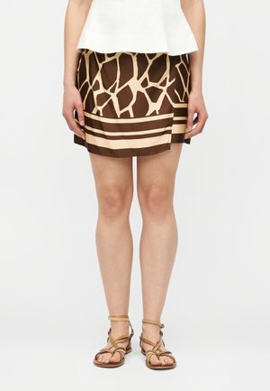 Femme portant un haut blanc, une mini-jupe à motifs marron et beige, et des sandales plates à lanières couleur camel, debout devant un fond uni.