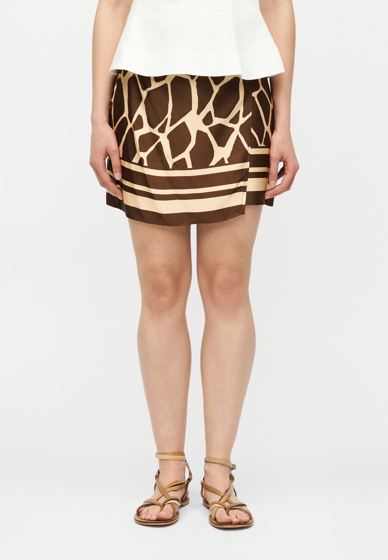 Γυναίκα που φοράει λευκή μπλούζα, καφέ και μπεζ μίνι φούστα με σχέδια, και ταμπά πλατφόρμες με λουράκια, στέκεται μπροστά σε απλό φόντο.