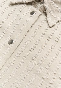 Beige Denimhemd mit strukturierten Streifen, ausgefransten Details und metallischen Druckknöpfen, ausgestattet mit einem spitzen Kragen und weichem Stofffinish.