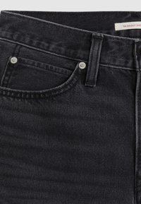 Détail d’un jean en denim noir montrant la poche avant, la boucle de ceinture et une partie d’une étiquette indiquant "'94 BAGGY SHO."