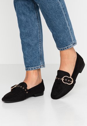 Fødder iført sorte loafers i ruskind med guldbeklædning, kombineret med blå opsmøgede jeans mod en ensartet lys baggrund.
