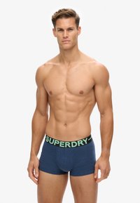 Męskie niebieskie bokserki wykonane z miękkiego materiału. Posiadają czarny pas z zielonym logo "SUPERDRY" i dopasowany krój.