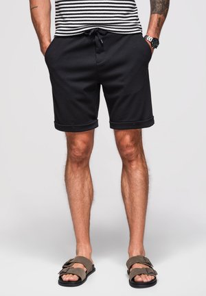 Mand iført sorte rullede shorts, stribet skjorte, ur og brune sandaler, der står med hænderne i lommerne på grå baggrund.