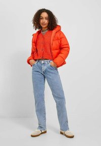 Giacca a vento arancione brillante con cappuccio, cardigan lavorato a maglia arancione, jeans blu chiaro a gamba dritta e scarpe beige con suola marrone.