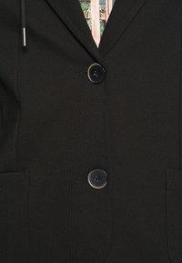 Blazer noir à deux boutons noirs, col cranté et poches plaquées, porté sur une chemise à motifs multicolores.