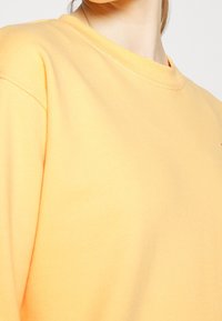 Sudadera amarilla con cuello redondo, hecha de algodón suave. Presenta mangas montadas y costuras visibles a lo largo de los bordes.