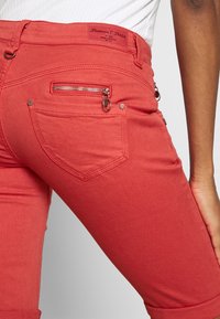 Punaiset puuvillasekoitefarkut, joissa istuva malli. Takana kaareva tasku ja sivussa vetoketjut yksityiskohtina toiminnallisuuden lisäämiseksi.