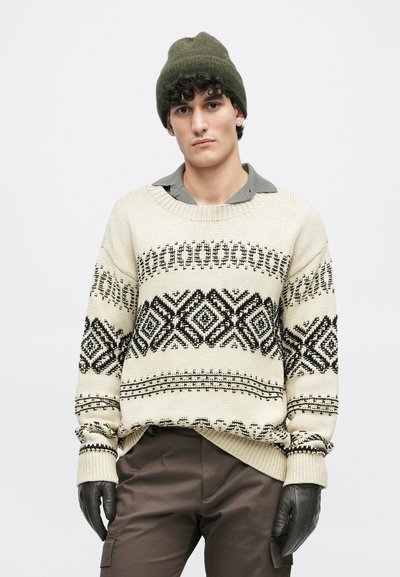 Krém színű kötött pulóver fekete geometrikus mintákkal, kerek nyakkivágással és bordás mandzsettával. Szürke galléros inggel és kesztyűvel viselve.
