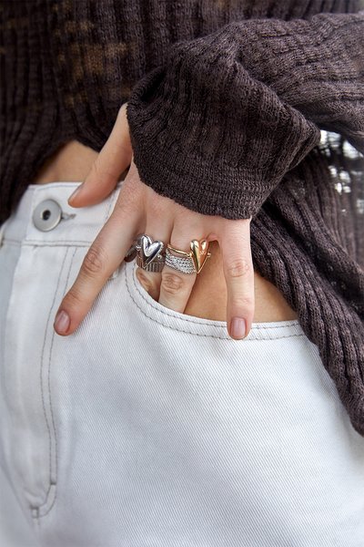 Drei Ringe an den Fingern: ein silberner Herzring, ein goldener Bandring mit einem Herz-Akzent und ein silberner Bandring mit klaren Steinen. Ein brauner Strickpullover ist sichtbar.