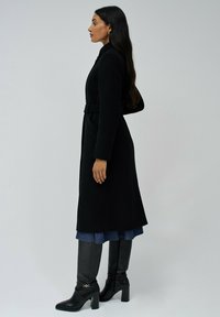 Zwarte wollen jas met een getailleerde pasvorm, lange mouwen, een riem en zijzakken. Combinatie met kniehoog zwarte laarzen en een denim rok.