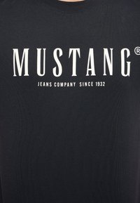 Melna kokvilnas T-kreklā ar lieliem baltajiem uzrakstiem: "MUSTANG" un "JEANS COMPANY SINCE 1932." Vienkāršs, arapa isu dizains ar ribotu tekstūru.