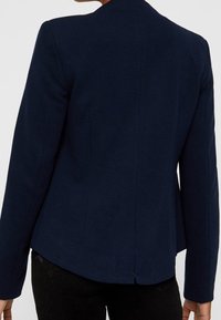 Blazer structuré bleu marine avec une finition texturée, des manches longues, un col haut et un ourlet légèrement courbé à l'arrière.