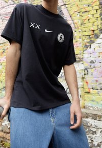 Camiseta de algodón negra con acentos gráficos blancos, incluyendo dos marcas de cruz y un logotipo de baloncesto, combinada con pantalones cortos de mezclilla azules.