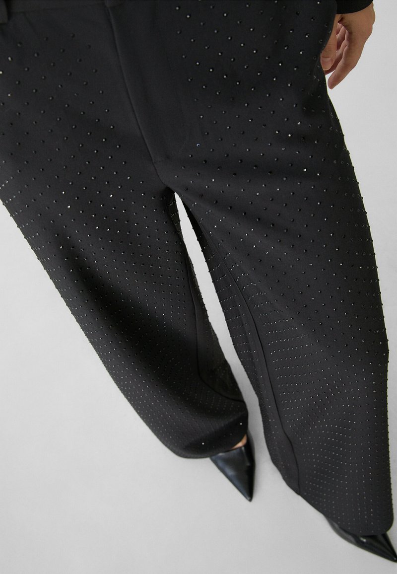 Pantalones de pierna ancha negros con pequeños adornos brillantes espaciados uniformemente, llevados con zapatos negros de punta, mostrando parte de una mano.