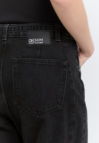 Vaqueros de mezclilla negra con un acabado texturizado, costuras visibles y una etiqueta. Presenta un bolsillo trasero con un ángulo y forma distintivos.