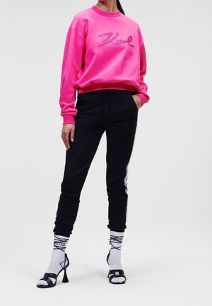Personne portant un sweat-shirt rose vif avec le texte "Karl", un pantalon de jogging noir avec une bande latérale, des chaussettes blanches avec logo, et des sandales noires à talons.